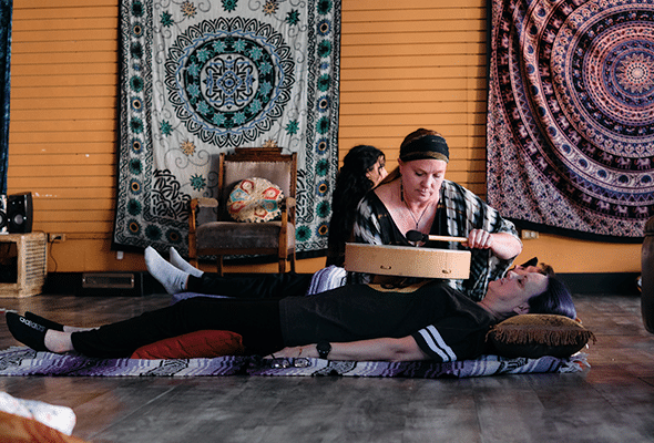 A woman lightly taps a drum atop another woman laying across the floor in a tapestry adorned eclectic room.