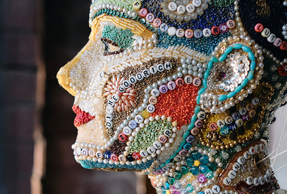 A mannequin head is beautifully adorned with beads, some spelling out names of iconic females through history.