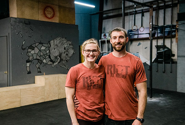 The male and female owners stand arm in arm at the front desk of Rebel Herd Fitness in Pittsburg, Kansas.