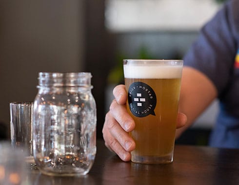 A full beer is set on a table with other glassware. The Brick and Mortar Social House logo is visible on the beer glass.