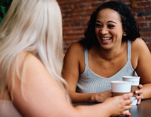 A woman laughs while sharing a cup of coffee with a friend.