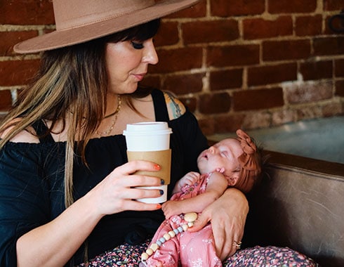 A young mother holds a newborn baby in one arm with a fresh cup of coffee in the other.