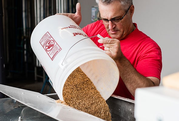 A man dumps dried grains into a large hopper to brew beer.