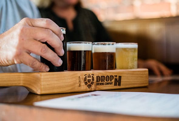 A wooden serving platter with a heat-branded logo of Drop the H brewing sits atop a table as a patron grabs a beer.