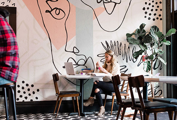 A woman sits in front of an abstract mural with a contour drawing of a face at Toast restaurant.