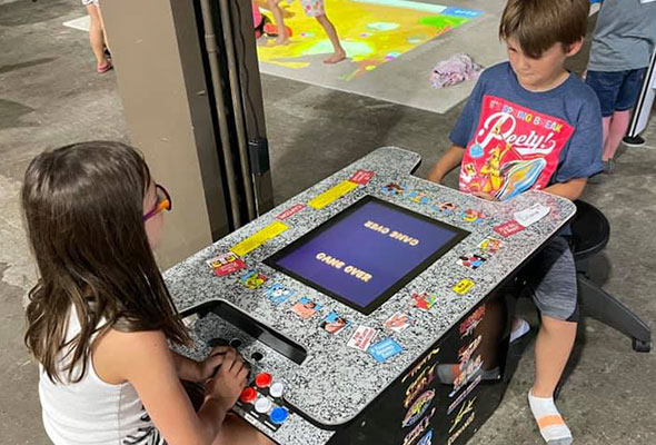 Two children sit across from one another at a sit-down cocktail arcade cabinet.