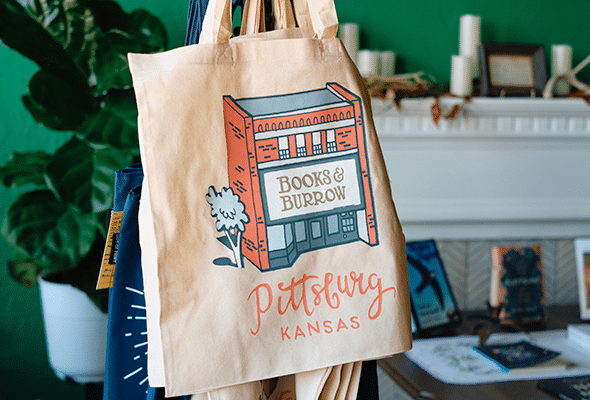 A shopping bag hangs on a rack showcasing a colorful illustration of Books & Burrow, highlighting its Pittsburg home.