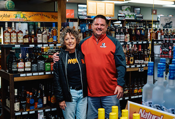 The mother and son owners of Pairott Head Liquor stand hand in hand smiling at the camera inside their store.