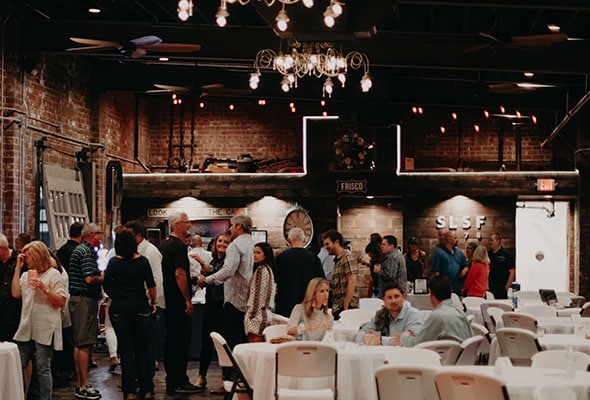 A large group converses amidst white table cloth adorned tables.